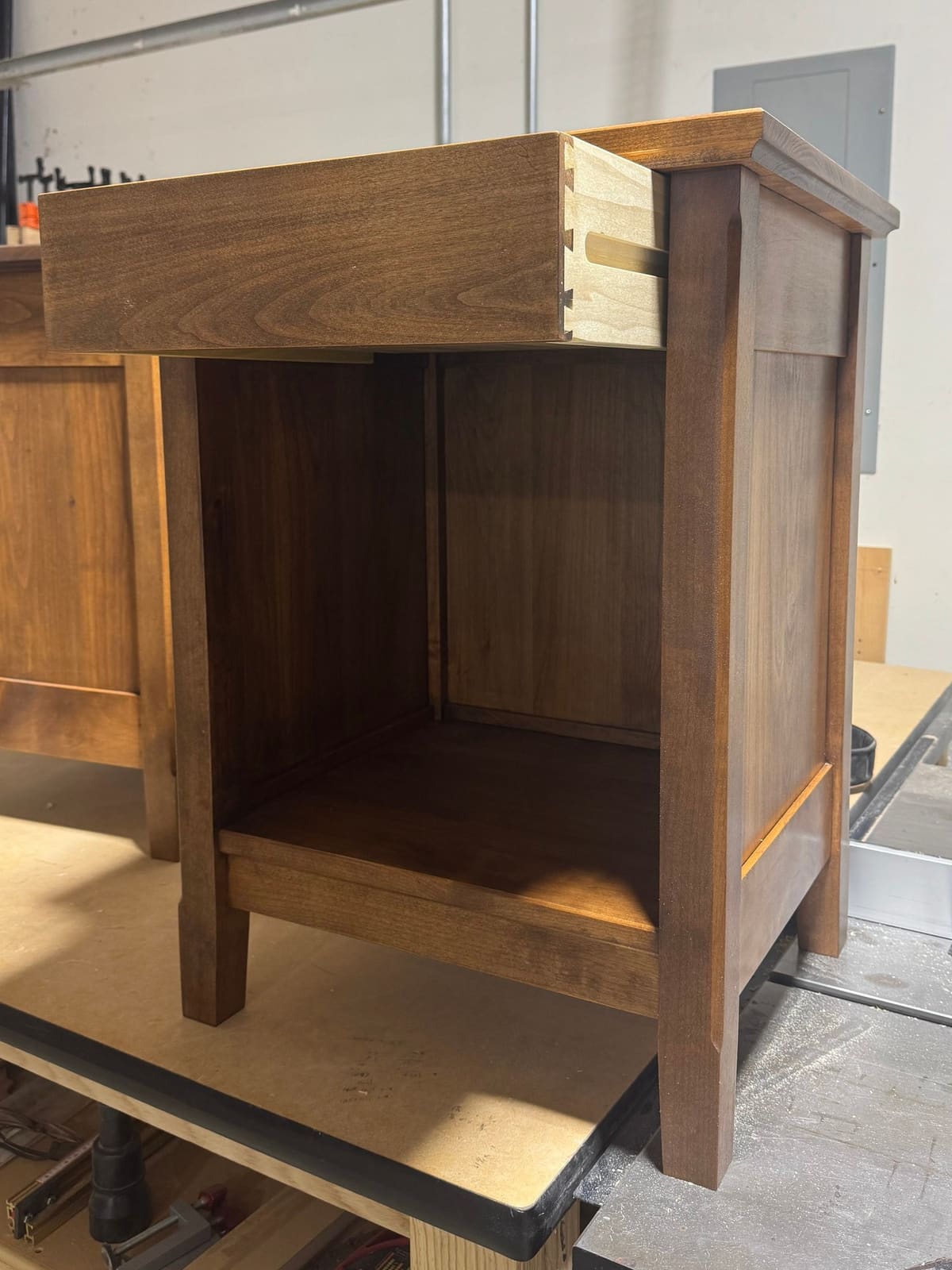 Stained nightstand on a shop bench with its drawer pulled out to show dovetailed corners
