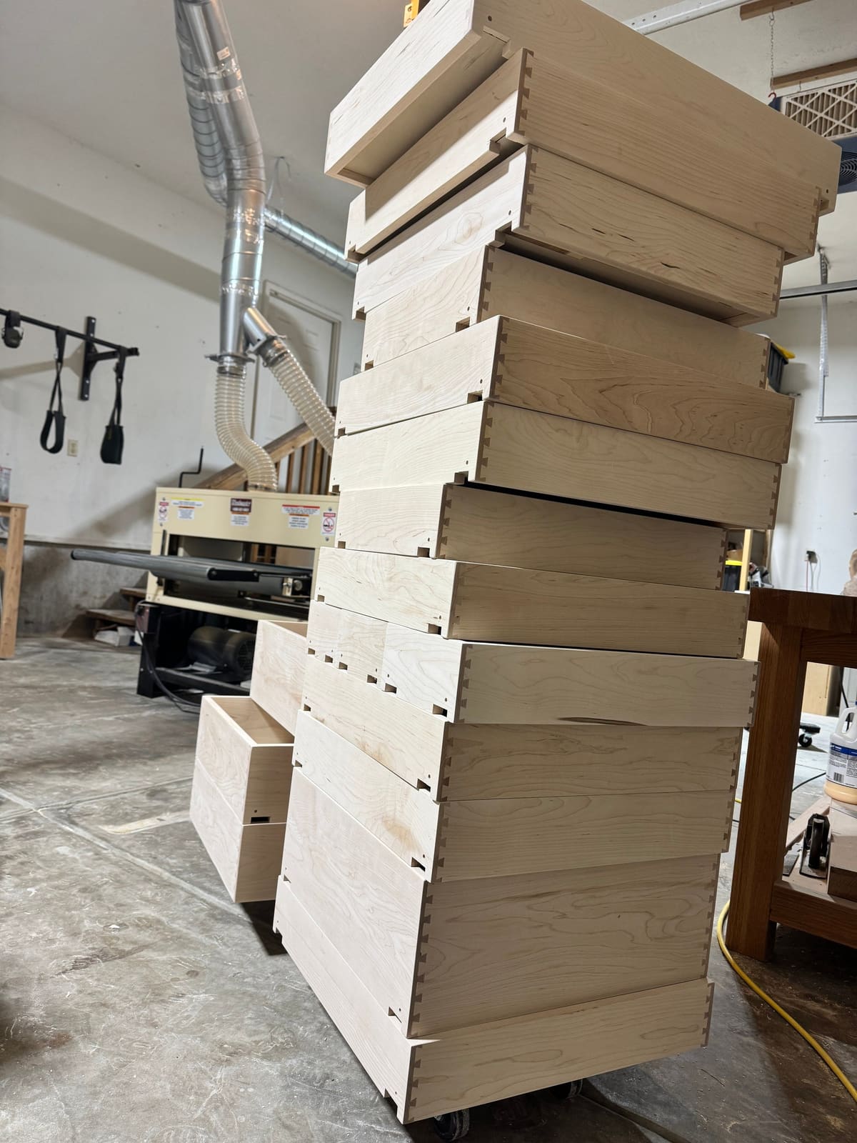 Stack of dovetailed drawer boxes in a light wood on a cart in a woodworking shop with dust collection ducts