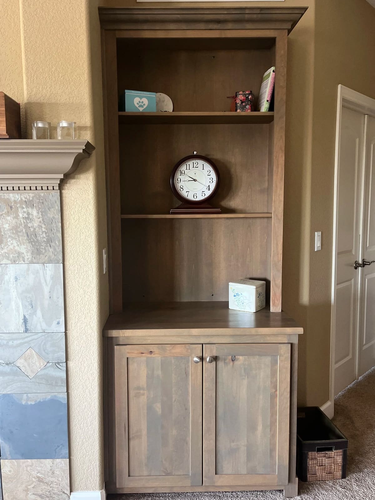 Gray-stained built-in bookcase with open shelves above a two-door shaker cabinet beside a tiled fireplace