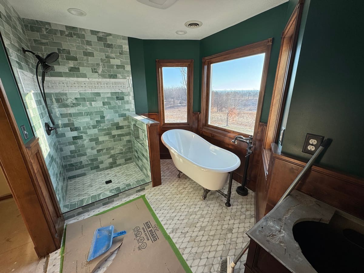 Bathroom with stained wood wainscoting and window casings around a clawfoot tub, next to a green tiled shower
