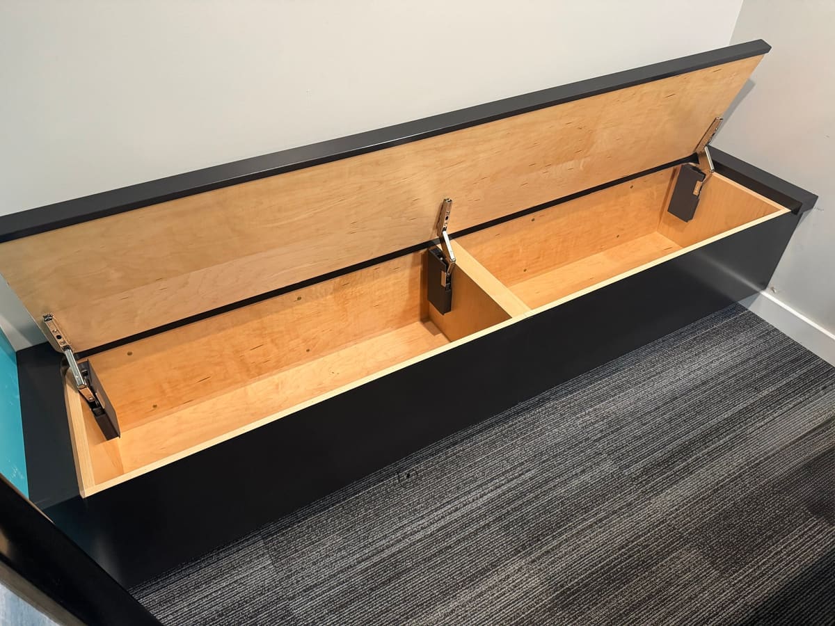 Black built-in storage bench with its lid open, showing a light wood interior with a center divider and lid stays