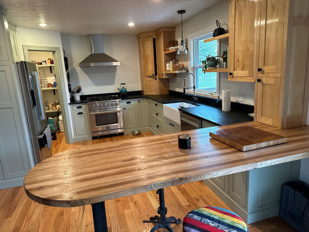 Kitchen with sage green lower cabinets, natural wood uppers, dark counters and a rounded butcher-block peninsula