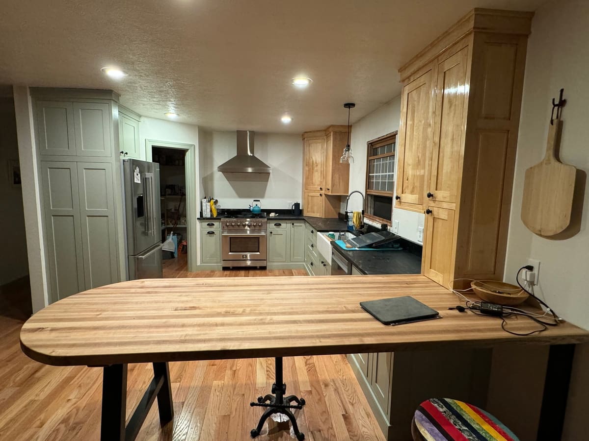 Evening view of a sage and natural wood kitchen across a butcher-block peninsula on black steel legs