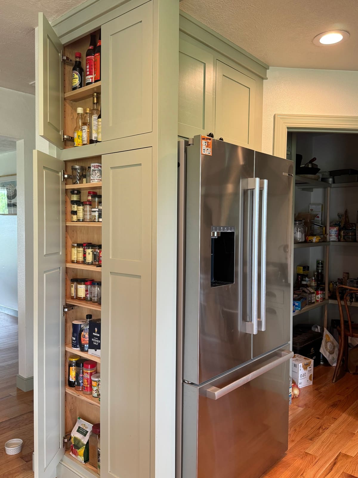 Tall sage green pantry cabinet with doors open to adjustable spice shelves, beside a refrigerator surround