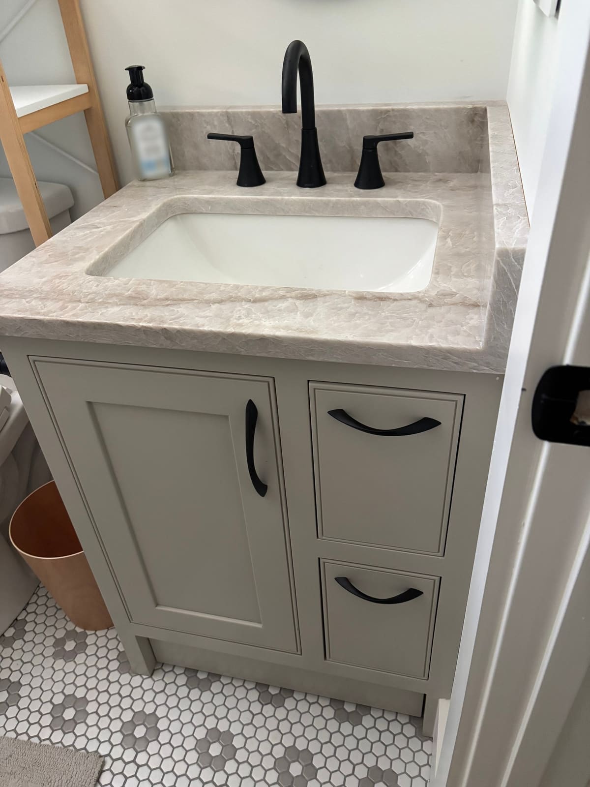 Greige inset bathroom vanity with one door, two drawers, black pulls, and a stone top with undermount sink