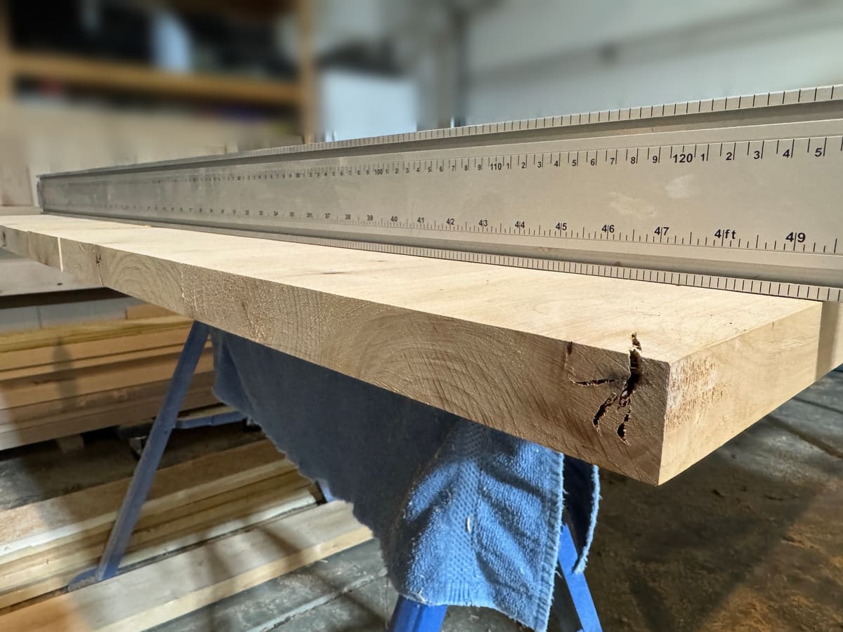 Thick hardwood plank on a sawhorse under a long aluminum straightedge in a woodworking shop