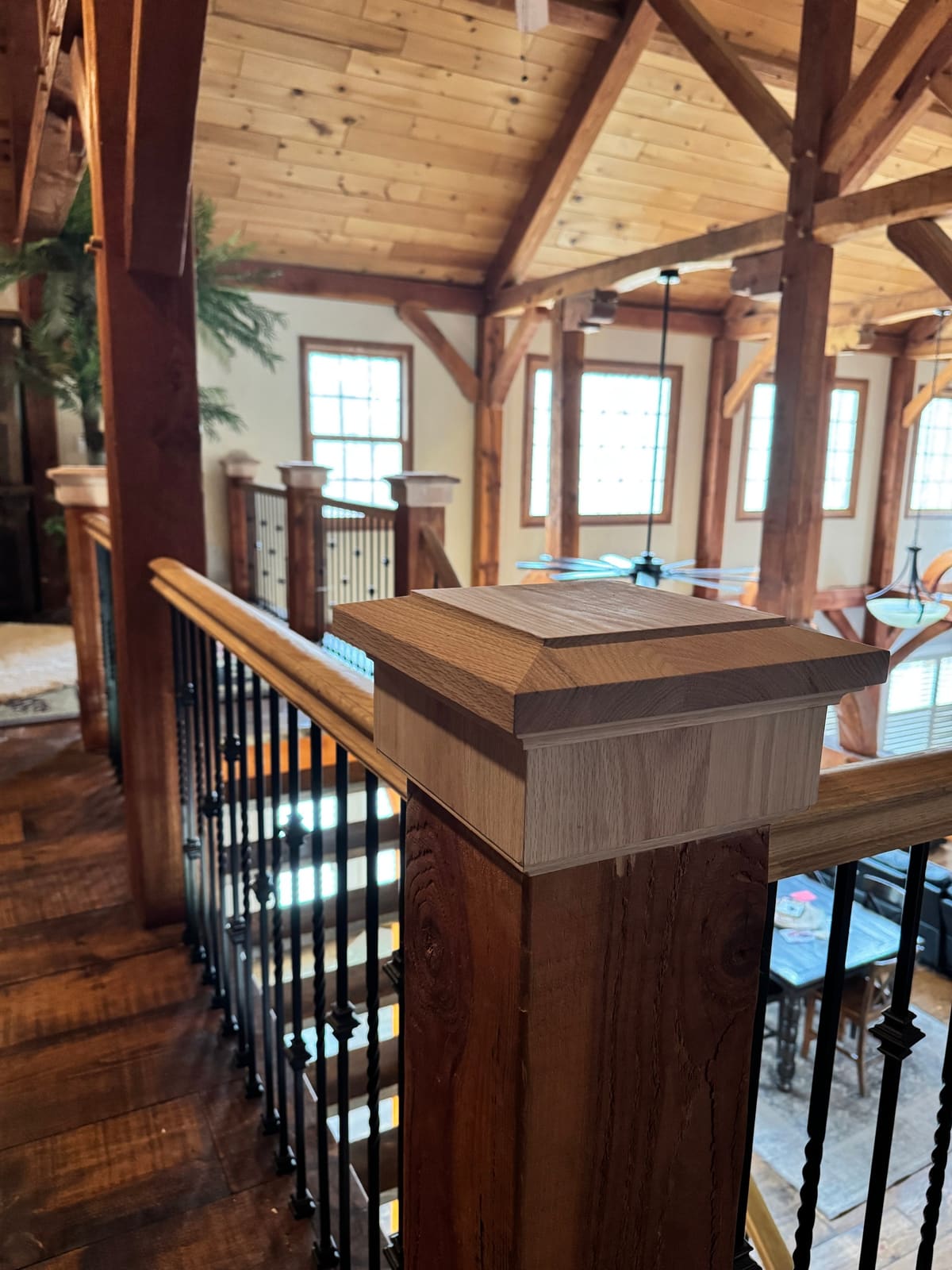 Stained newel post with a stepped, beveled light wood cap, handrail and twisted iron balusters in a timber-frame home