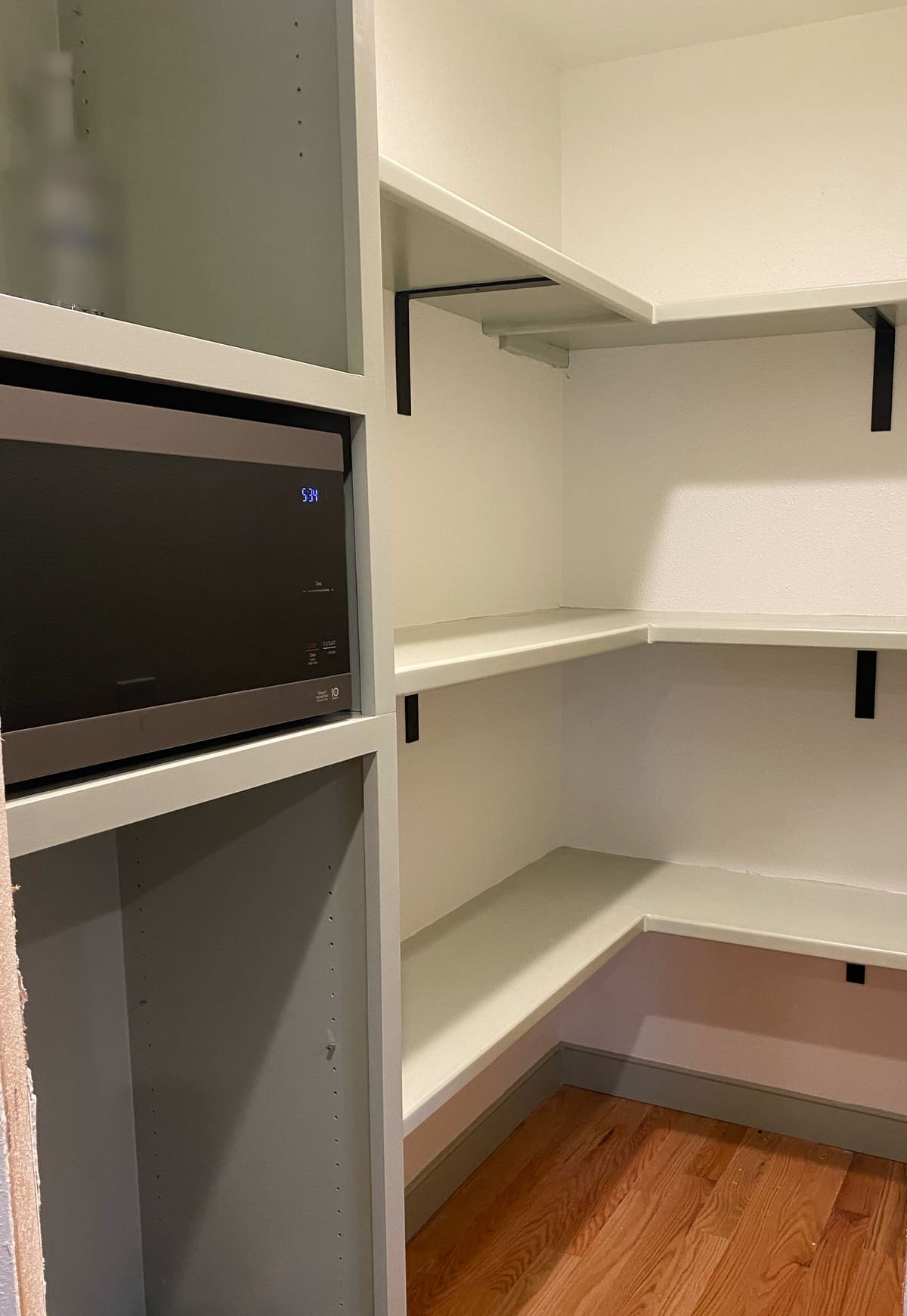 Walk-in pantry with painted wraparound shelves on black brackets and a tall sage cabinet with a built-in microwave